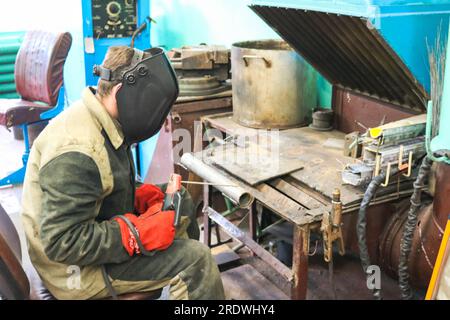 Un lavoratore maschio un saldatore in una maschera protettiva salda un tubo metallico in una stazione di saldatura in un'officina in un impianto metallurgico in un impianto di riparazione. Foto Stock