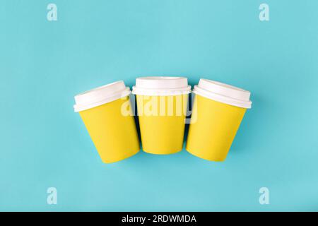 Tre tazze di carta gialla monouso con coperchi in plastica chiusi per caffè da asporto, che è poi riciclabile. Modello piatto vuoto poggiato su sfondo blu Foto Stock