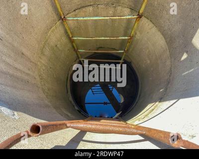 Un grande pozzo rotondo profondo realizzato con anelli di cemento con pozzo pulito di acqua sotterranea per bere con vecchie scale arrugginite. Foto Stock