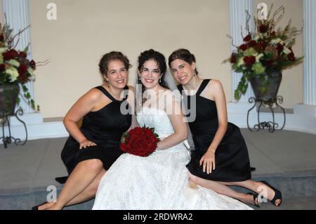 Sposa e le sue due sorelle siedono sui gradini della cappella nuziale. Due sorelle sono anche damigelle d'onore che indossano abiti neri lunghi al ginocchio. Sono tutti sorridenti Foto Stock