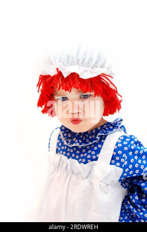 La bambina arrabbiata e incrociata è vestita con un costume da bambola da straccio. I suoi capelli sono in filato rosso ricoperti da un cappello bianco per mop. Foto Stock