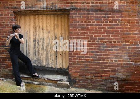 Musicista di strada donna, vestita di nero, si trova nella porta chiusa e sigillata di un vicolo e suona il suo flauto. Si sta appoggiando contro un muro di mattoni dentro Foto Stock