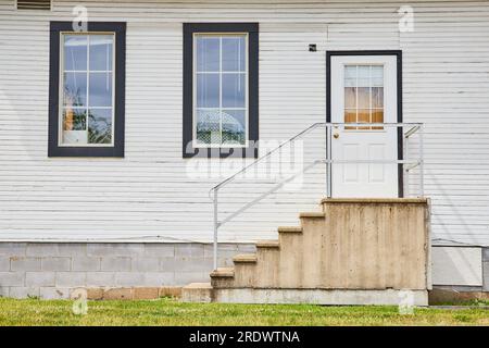Casa comune con porta bianca e scalini in cemento e oggetti che affollano le finestre esterne Foto Stock