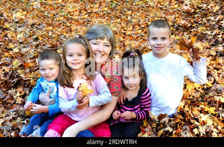 Rastrellare le foglie e divertirsi è la tradizione di questa famiglia. La nonna tiene due dei suoi nipoti e gli altri due si siedono accanto a lei. Sono bur Foto Stock