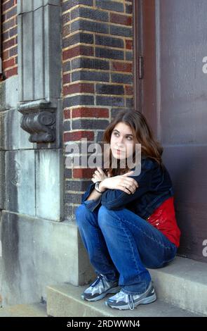 La giovane studentessa delle superiori si siede sui gradini della sua scuola e si perde nel pensiero. Indossa una giacca in denim e dei jeans. Foto Stock