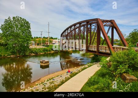 Persona seduta su un muro di cemento sotto un ponte di ferro con fiume che scorre in estate Foto Stock