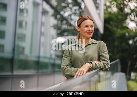 Una donna d'affari di mezza età si trova sullo sfondo di un moderno edificio per uffici e guarda di lato Foto Stock