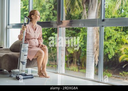 La giovane donna incinta ama pulire la sua casa. Facile pulizia con un aspirapolvere wireless. Donna incinta che pulisce il pavimento con l'aspirapolvere portatile Foto Stock