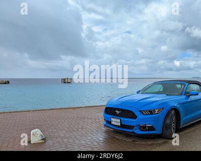 Un'auto Ford Mustang dal colore blu vivo si trova sul bagnato lungomare di Kralendijk, Bonaire, Leeward Antilles, con una vista sul verde e blu dei Caraibi. Foto Stock