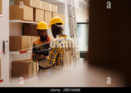 I dipendenti di Storehouse ascoltano le istruzioni del responsabile logistico sul telefono fisso e l'assistente prende appunti negli appunti. Due operai afroamericani che parlano al telefono con il responsabile delle spedizioni Foto Stock