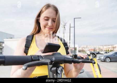 Donna sorridente che noleggia uno scooter elettrico a spinta tramite l'app per telefono cellulare Foto Stock