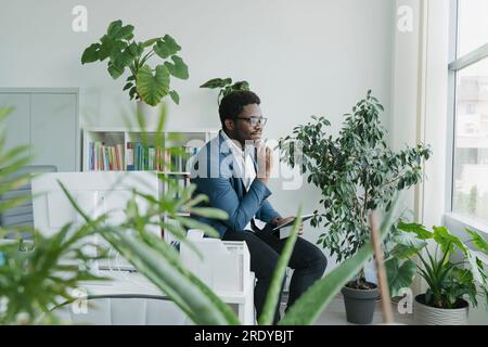 Un uomo d'affari premuroso seduto con un blocco note e guardandolo dalla finestra all'ufficio Foto Stock