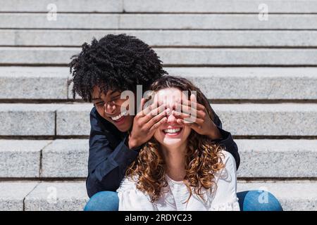 Allegra giovane donna che copre gli occhi di un amico seduto sulle scale Foto Stock