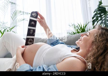 Donna incinta che guarda ecografie sdraiata sul divano a casa Foto Stock