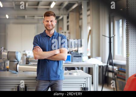 Tecnico sorridente con le braccia incrociate in piedi accanto al tavolo nell'industria Foto Stock