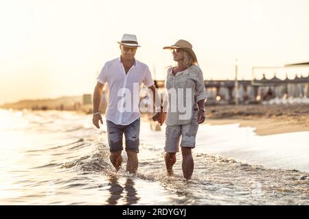 Una coppia di mezza età che cammina mano nella mano sulla spiaggia al tramonto. Foto Stock