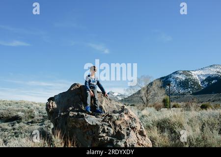 Il ragazzo che indossa gli occhiali da sole si siede su enormi montagne innevate di roccia sullo sfondo Foto Stock