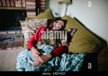 Un uomo che dorme su un divano con un gatto marrone che dorme su di lui Foto Stock