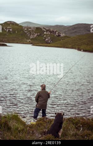 uomo e cane sono seduti a pesca con la mosca sul lago nelle remote montagne della scozia Foto Stock
