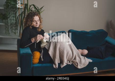 Giovane donna con i capelli ricci lavorati a maglia mentre si gode un weekend confortevole a casa. Attività hobby preferita nel weekend, concetto di benessere Foto Stock