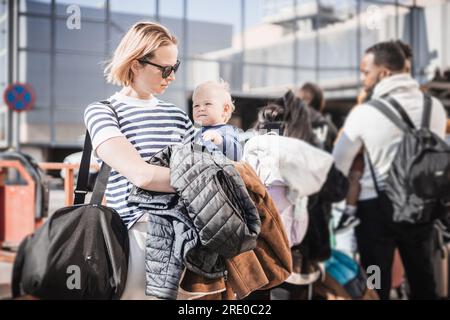 La madre viaggia con il suo bambino. La mamma tiene in mano la borsa da viaggio e il suo bambino mentre fa la fila per l'autobus di fronte alla stazione del terminal dell'aeroporto. Foto Stock