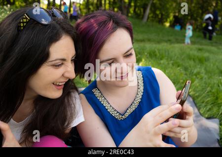 Due giovani donne, amici o sorelle che guardano qualcosa al telefono, ridono, si rilassano in un parco naturale. Foto Stock