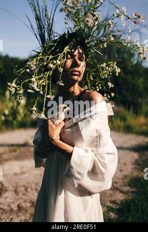 Una giovane donna con un vestito bianco e una corona di margherite tiene in mano un piccolo chiken, sullo sfondo della foresta, la luce del tramonto Foto Stock