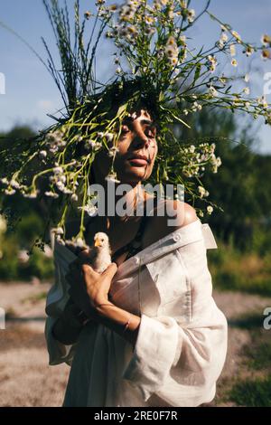 Una giovane donna con un vestito bianco e una corona di margherite tiene in mano un piccolo chiken, sullo sfondo della foresta, la luce del tramonto Foto Stock