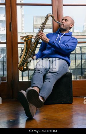 giovane latinoamericano di colore blu, seduto in salotto a casa, molto ispirato ed eccitato a suonare il sassofono, a fare pratica e a fare musica per passare il Foto Stock