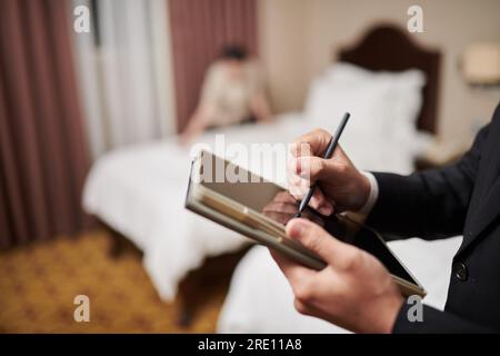 Immagine ravvicinata del manager dell'hotel che compila il modulo sul tabulato lascia che il computer quando la cameriera fa il letto sullo sfondo Foto Stock