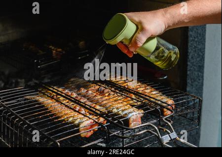 Il pesce viene cotto su una griglia metallica in una fiamma di fuoco. La mano cosparge di olio d'oliva, spezie di pesce, cotto alla griglia. Foto Stock