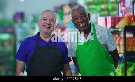 Lavoratori anziani soddisfatti e diversificati che si trovano in un negozio di alimentari sorridendo davanti alla macchina fotografica indossando uniformi. Lavoratori del personale brasiliano più anziani Foto Stock