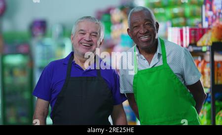 Lavoratori anziani soddisfatti e diversificati che si trovano in un negozio di alimentari sorridendo davanti alla macchina fotografica indossando uniformi. Lavoratori del personale brasiliano più anziani Foto Stock