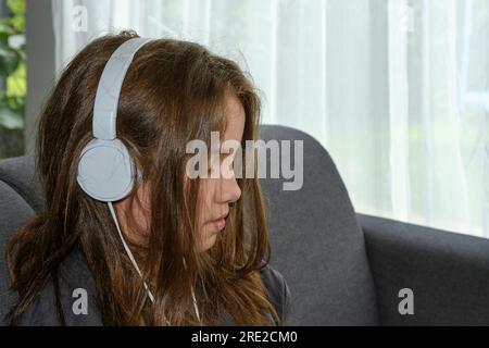 La giovane ragazza con le cuffie a casa seduta sul divano usa un tablet sullo sfondo di una finestra. Stile di vita Foto Stock