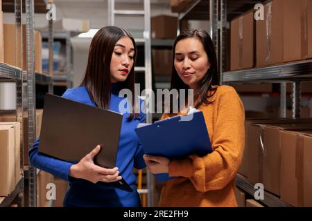 Dipendenti di donne asiatiche che gestiscono le scorte e la distribuzione dei prodotti. I responsabili di magazzino controllano le merci nello storage, eseguono l'inventario utilizzando i dati presenti negli appunti e nei laptop Foto Stock