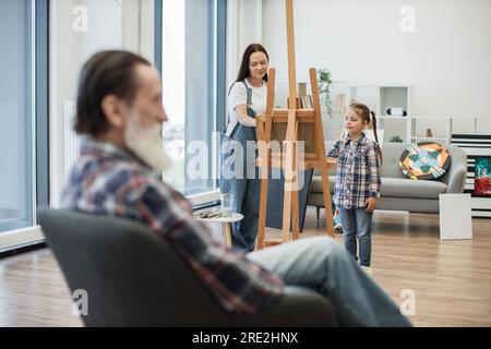 Attenzione selettiva delle donne caucasiche con abiti in denim che dipingono su tela, mentre un uomo più anziano posa in sedia in primo piano. Madre e figlia d ispirate Foto Stock