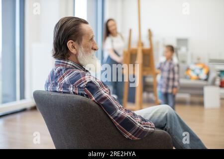 Vista posteriore di un signore anziano che riposa in una stanza luminosa mentre la donna e la ragazza caucasiche sono impegnate nella pittura. Adulto anziano rilassato che assiste il piccolo gr Foto Stock