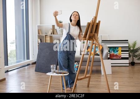 Giovane bella femmina sorridente mentre fai selfie su gadget personali in un elegante appartamento con grandi finestre panoramiche. Artista creativo che posa vicino a un grande cavalletto di legno e a un piccolo tavolo con pitture. Foto Stock