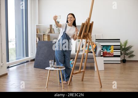 Giovane bella femmina sorridente mentre fai selfie su gadget personali in un elegante appartamento con grandi finestre panoramiche. Artista creativo che posa vicino a un grande cavalletto di legno e a un piccolo tavolo con pitture. Foto Stock