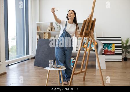 Giovane bella femmina sorridente mentre fai selfie su gadget personali in un elegante appartamento con grandi finestre panoramiche. Artista creativo che posa vicino a un grande cavalletto di legno e a un piccolo tavolo con pitture. Foto Stock