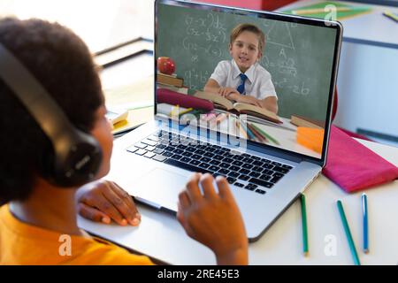 Vista posteriore di un ragazzo birazziale che ha una videochiamata con un ragazzo caucasico sul computer portatile a scuola Foto Stock