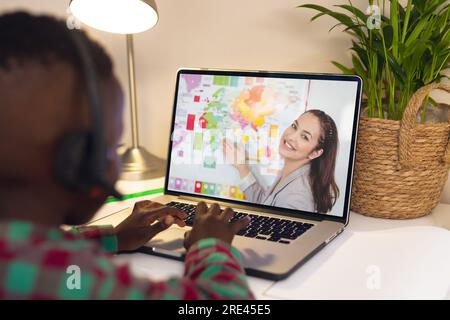 Vista posteriore di un ragazzo afro-americano che utilizza un computer portatile mentre effettua una videochiamata con un'insegnante birazziale Foto Stock