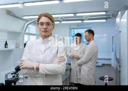 Ritratto di donna scienziata matura seria, capo di ricerca leader del gruppo in piedi all'interno del laboratorio indossando cappotto medico bianco e occhiali protettivi e guardando la macchina fotografica. Foto Stock