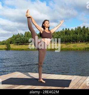 Praticante di yoga donna, esegue esercizi di Utthita Hasta Padangushthasana, equilibrio su una gamba, si allena con abbigliamento sportivo in piedi su un ponte di legno sui laghi Foto Stock