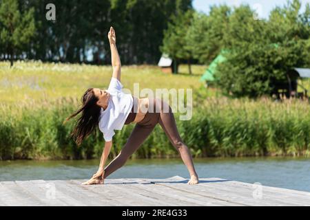 Una donna che conduce uno stile di vita sano e pratica yoga, esegue l'esercizio di Utthita Trikonasana, una posa triangolare, si allena in abbigliamento sportivo su una sunn Foto Stock
