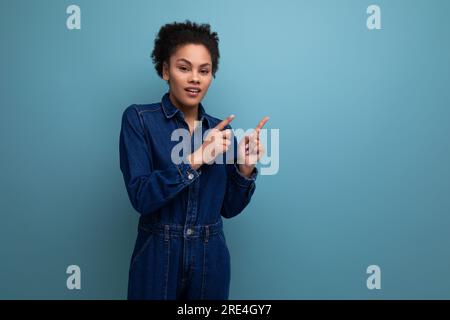 la giovane donna bruna ispanica, sottile e sottile, con morbidi capelli ricci e un abito in denim blu, si muove attivamente con le mani Foto Stock