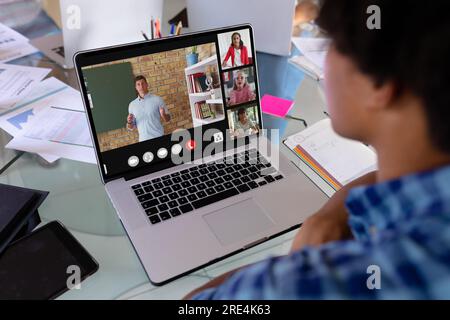 Vista posteriore di un ragazzo birazziale che tiene una videoconferenza con diversi studenti sul computer portatile a casa Foto Stock