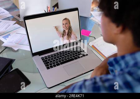 Vista posteriore di un ragazzo birazziale che ha videochiamato con una donna caucasica che insegna sul computer portatile a casa Foto Stock