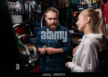 Riparatore professionista di ciclismo di moda con barba che comunica con una bella cliente bionda donna, che parla di problema di bicicletta, rilevato durante Foto Stock