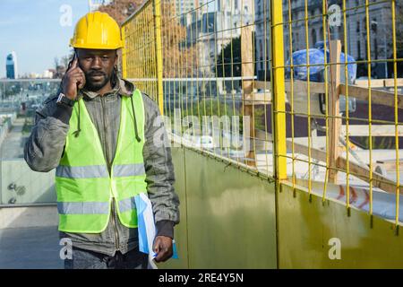 giovane ingegnere di etnia africana, supervisore che parla al telefono e cammina per il cantiere con una cartella in mano, indossa un hel Foto Stock